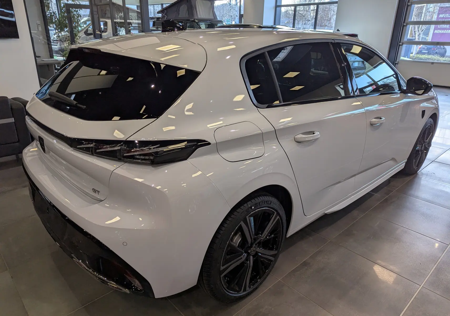 Peugeot 308 GT blanc Okénite vue 3/4 arrière droit en intérieur, avec jantes noires et vitres teintées.