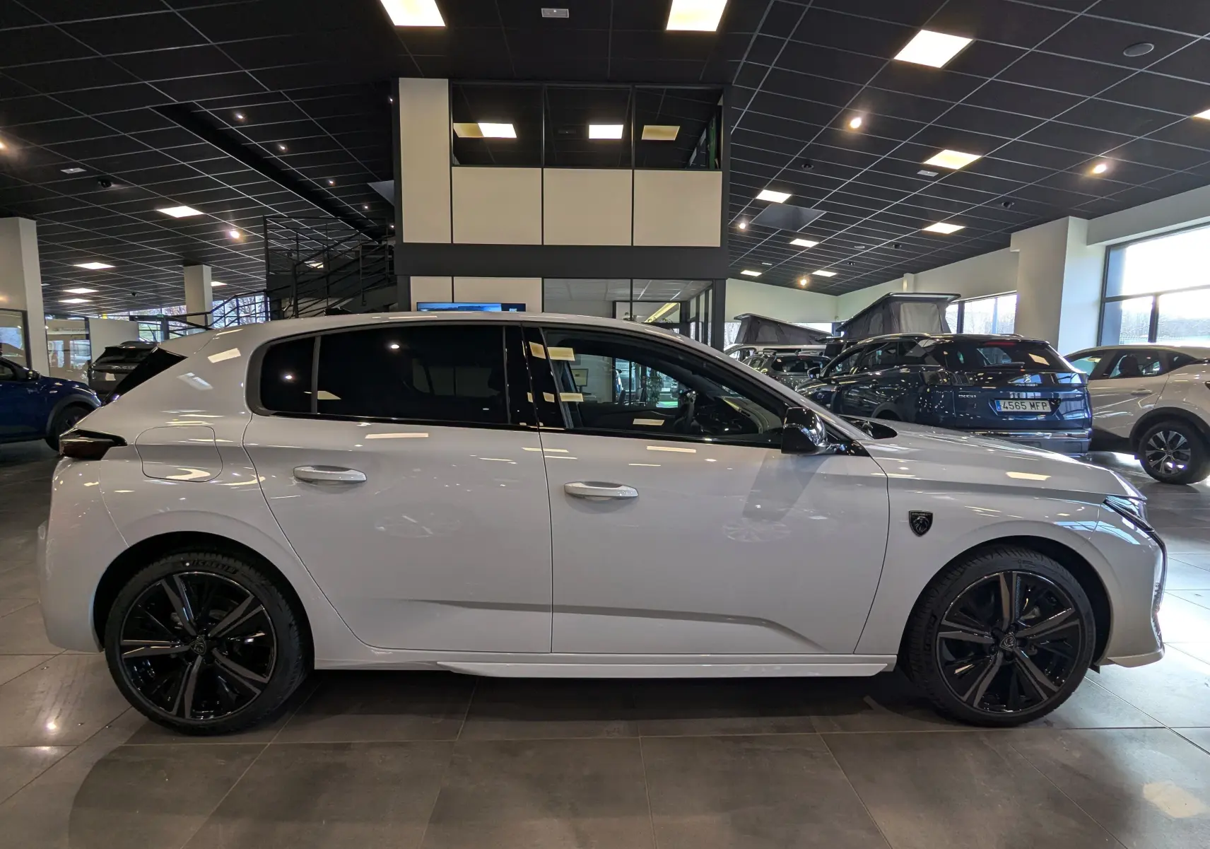 Profil latéral droit d'une Peugeot 308 GT blanche Okénite avec jantes noires dans un showroom lumineux.