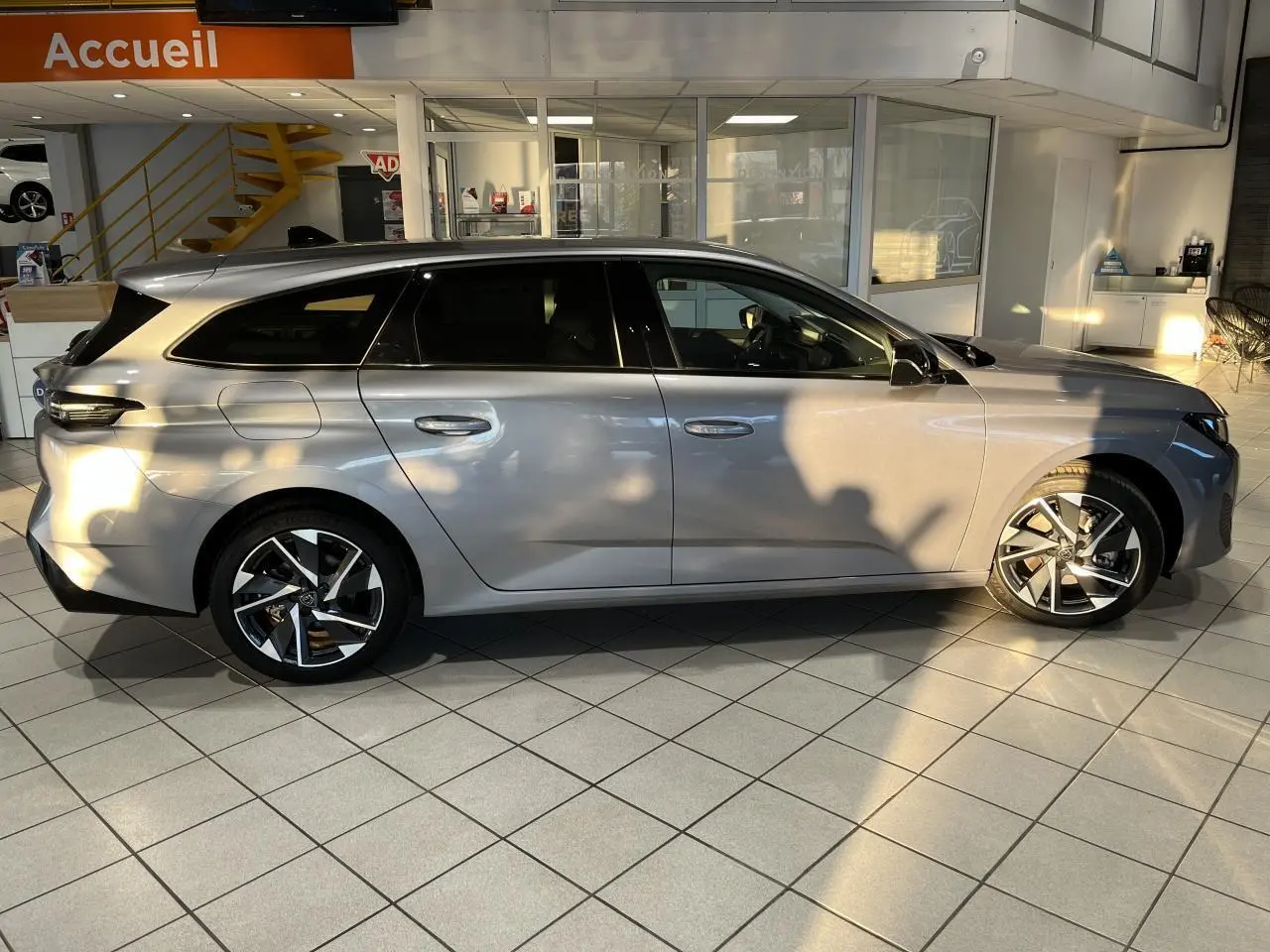 Peugeot 308 SW 2024 gris clair métal Artense vue de profil côté gauche en intérieur avec jantes alliage distinctives.