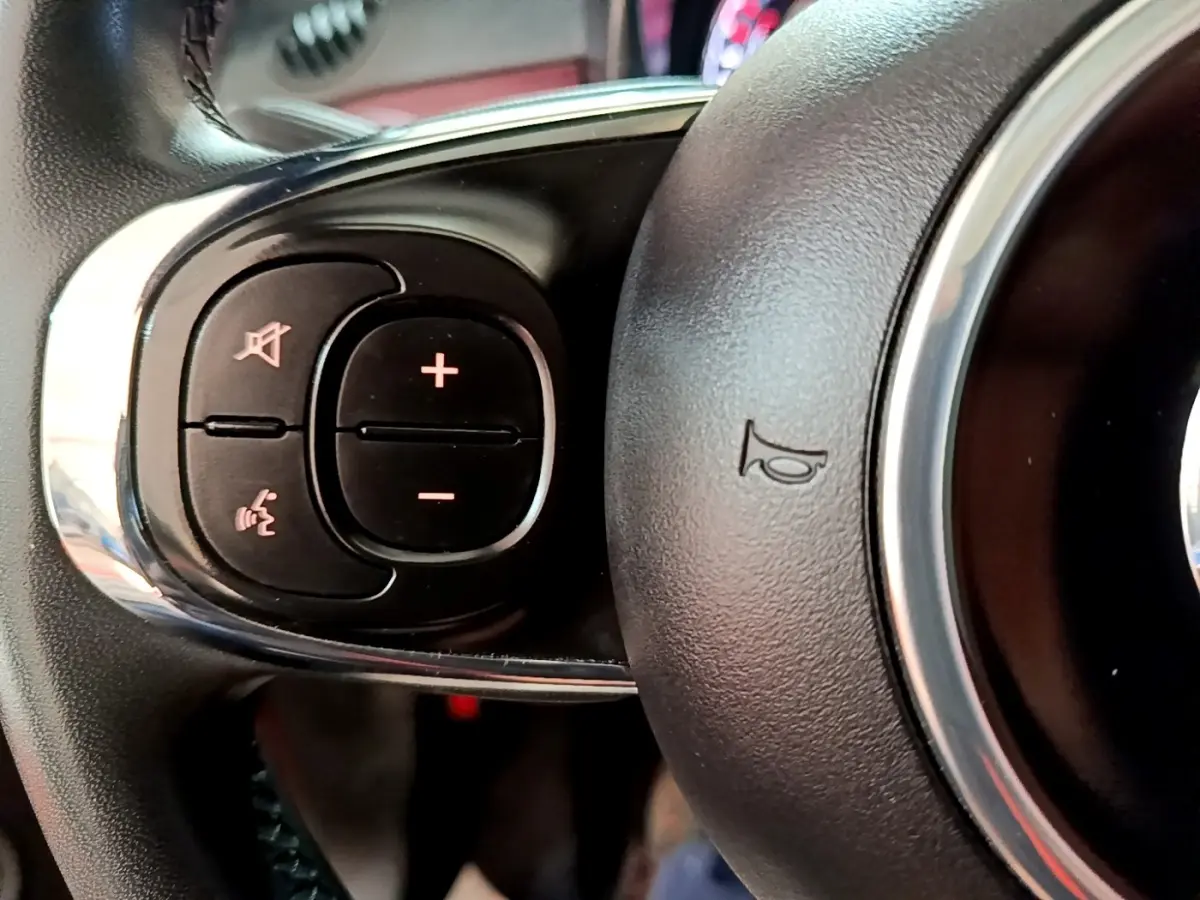 Gros plan sur les commandes gauche du volant noir de la FIAT 500 rouge, avec boutons de volume et reconnaissance vocale.