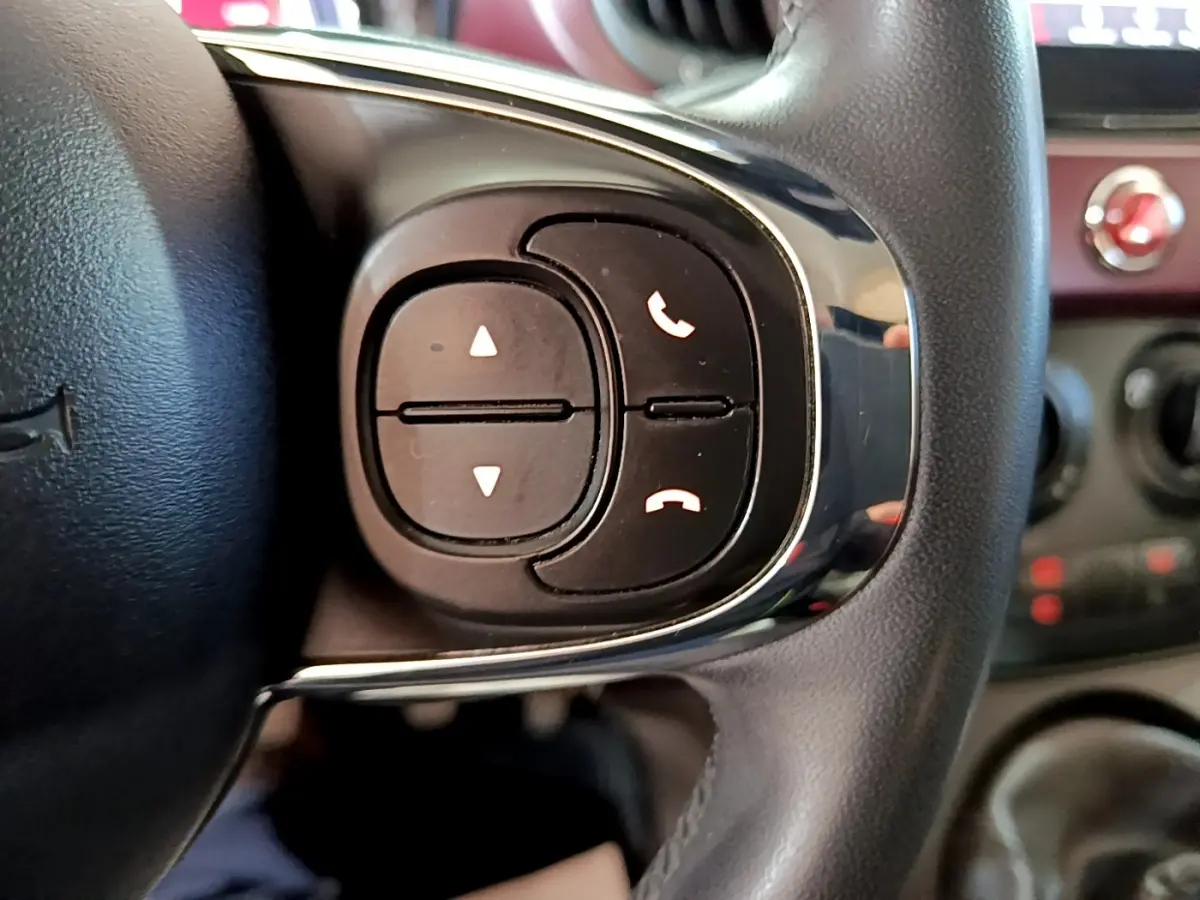 Gros plan sur les commandes noires du volant cuir de la FIAT 500 rouge, avec boutons de téléphone et réglages audio.