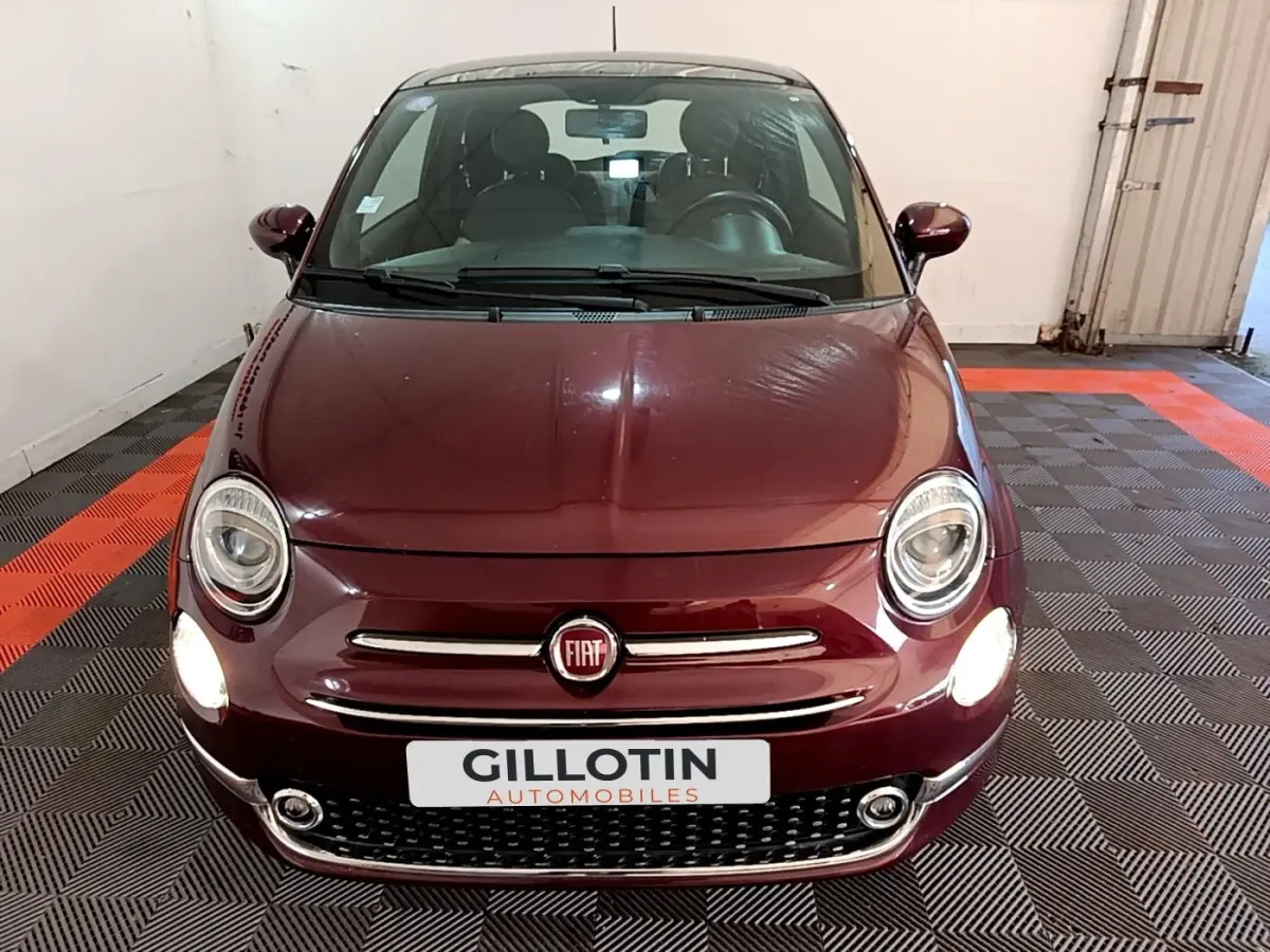 Vue avant d'une FIAT 500 rouge bordeaux avec phares allumés dans un garage au sol noir et rouge.