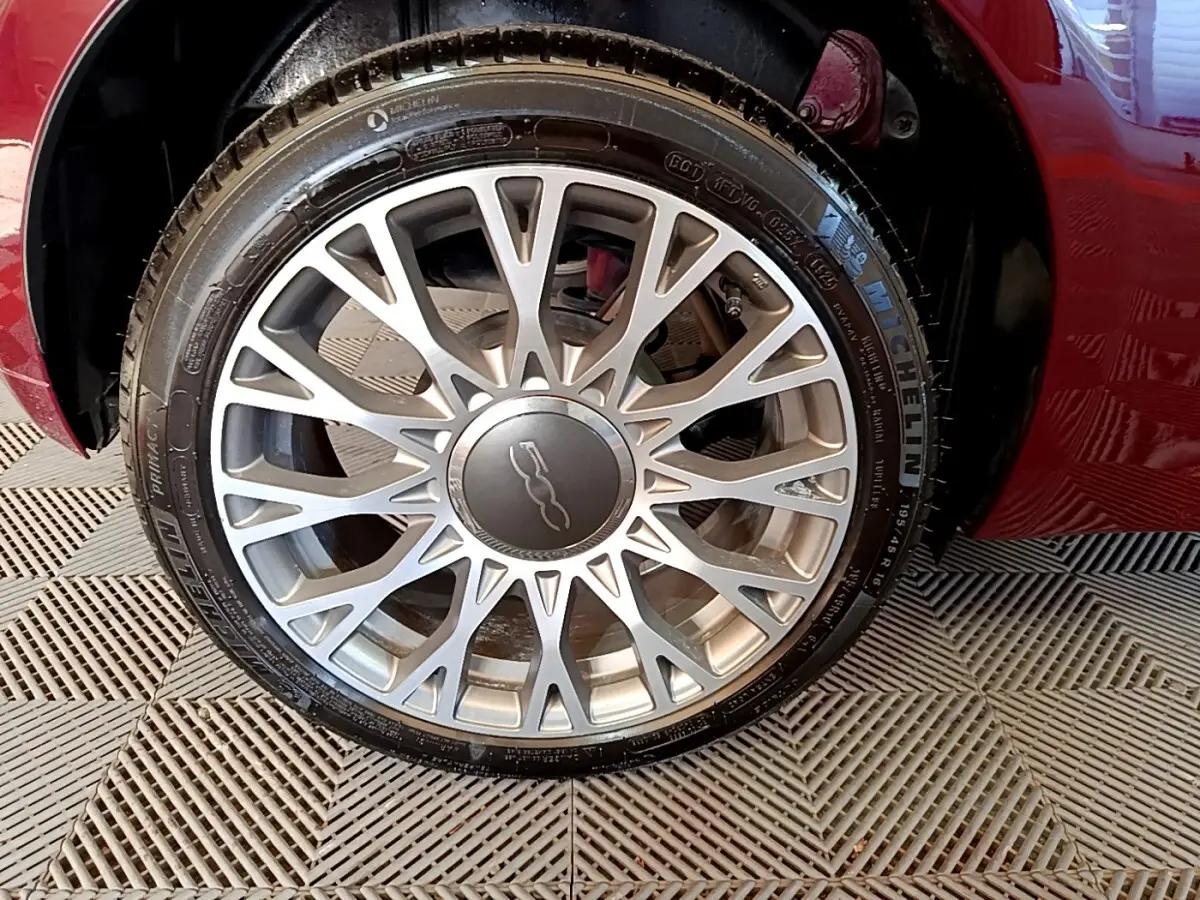 Gros plan sur la roue avant droite d'une FIAT 500 rouge, mettant en valeur la jante alliage argentée au design complexe.