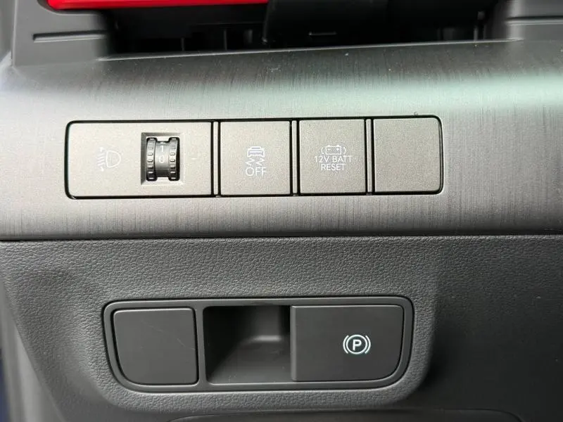 Gros plan sur les commandes grises du tableau de bord du Hyundai Kona 2025, avec réglage des phares et frein de parking électronique.