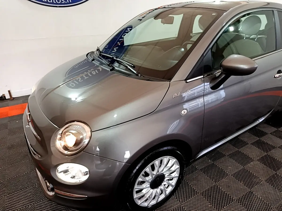 Vue 3/4 avant droit d'une FIAT 500 Dolcevita gris foncé avec phares allumés et jantes argentées détaillées.