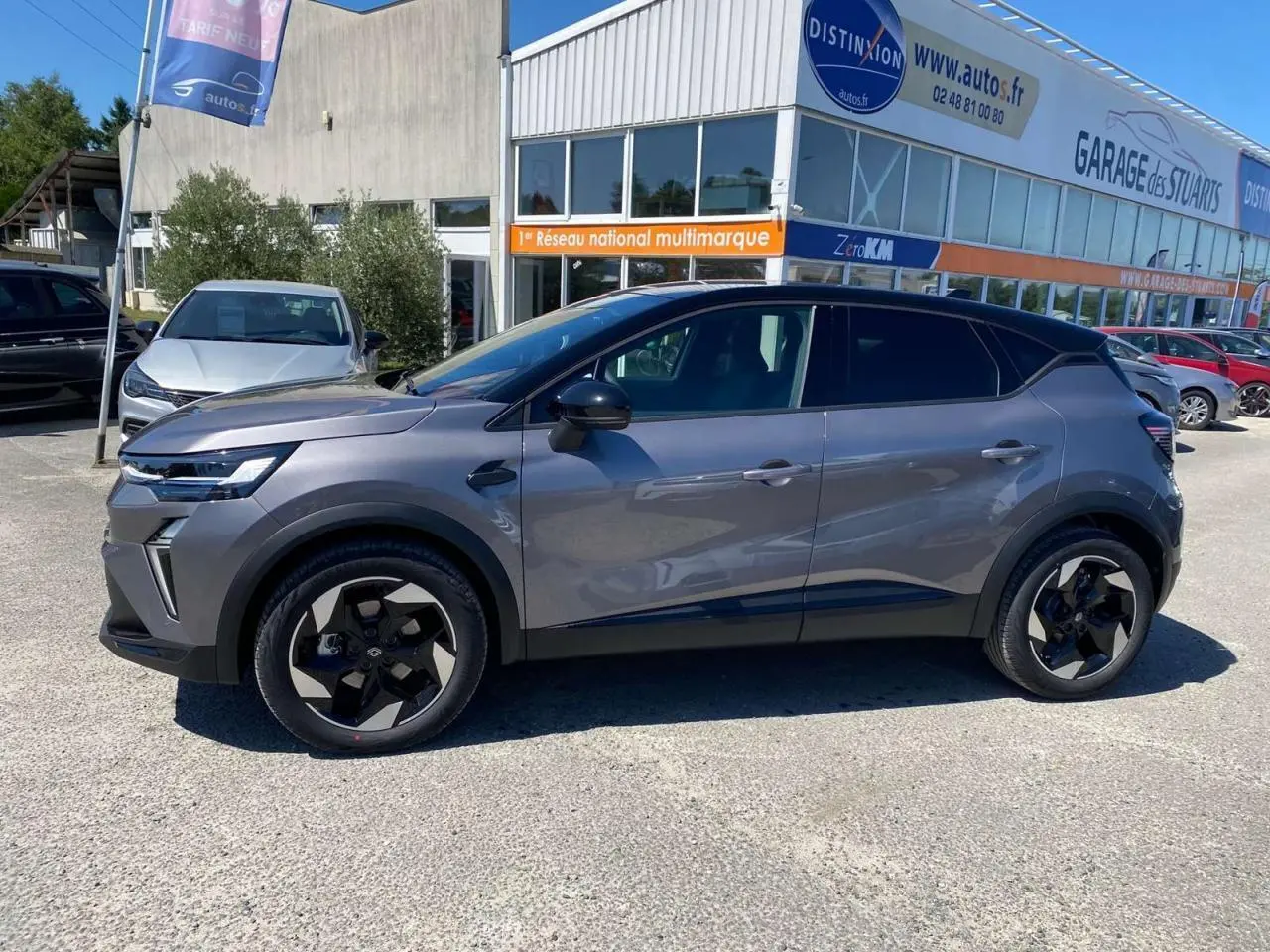 Profil côté gauche du Renault Captur gris Cassiopée avec toit noir, garé devant un garage multimarque.