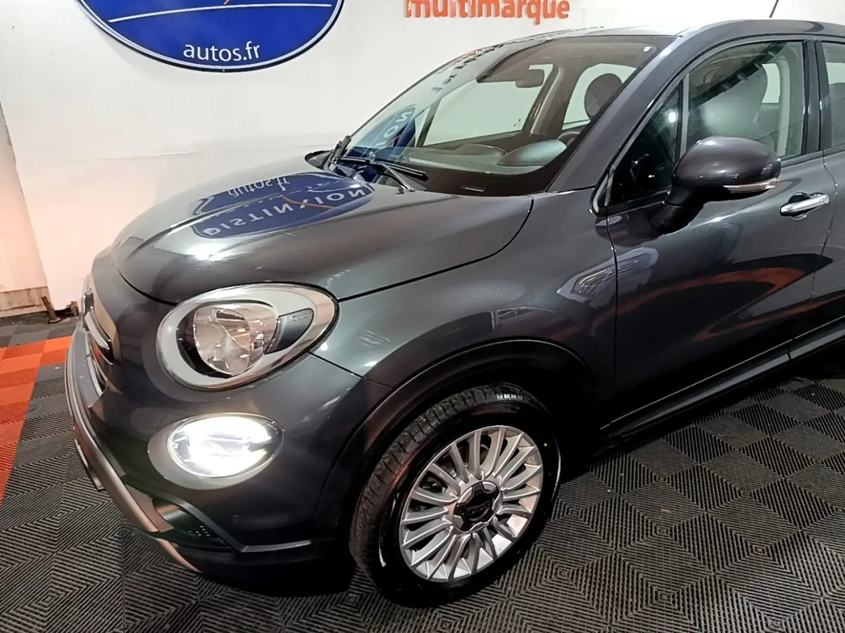 Vue 3/4 avant droit d'une FIAT 500X City Cross gris foncé avec phares et feux diurnes allumés en intérieur showroom.