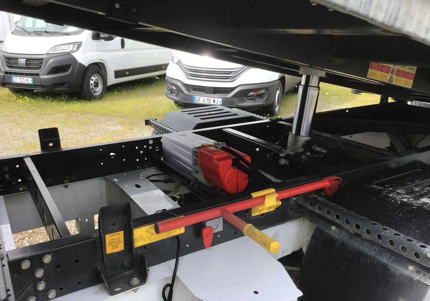 Vue rapprochée sous la benne du Renault Master blanc, montrant le châssis, vérin hydraulique et coffre de rangement.