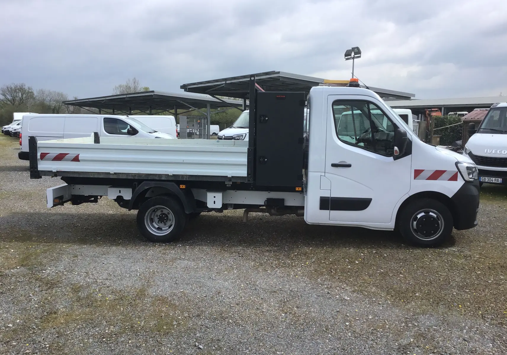Vue latérale droite d’un Renault Master blanc 2023 avec benne, coffre noir et roues jumelées sur terrain extérieur.