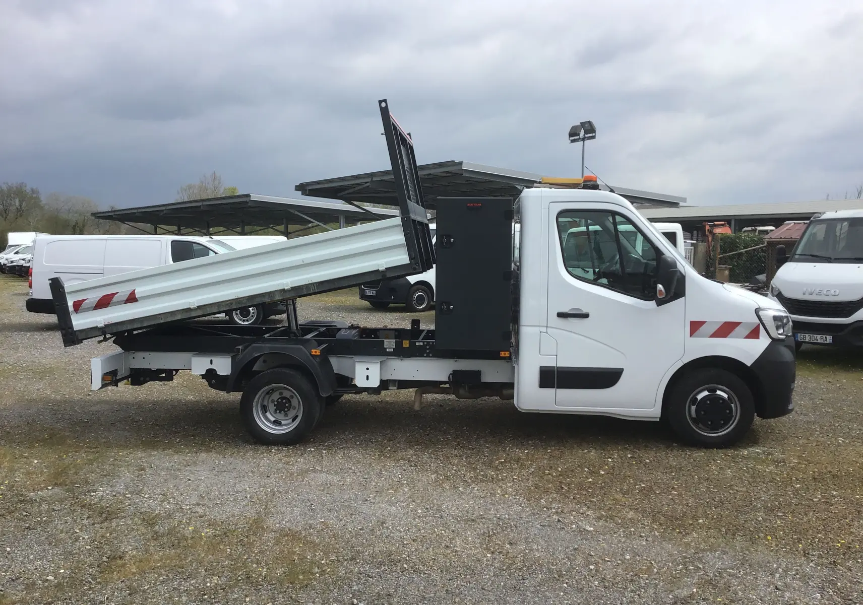 Vue latérale droite d’un Renault Master blanc 2023 avec benne relevée et coffre noir, roues jumelées visibles.