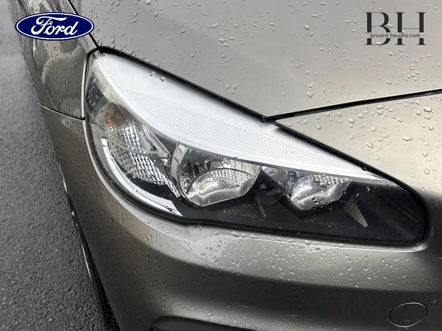 Gros plan sur le phare avant gauche d’une BMW Série 2 Active Tourer platinumsilber avec gouttes de pluie sur la carrosserie.