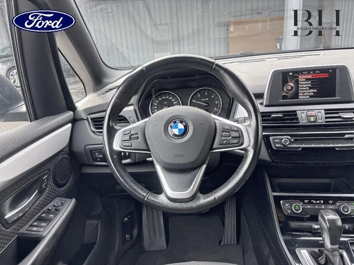 Vue intérieure centrée sur le volant cuir BMW et tableau de bord noir avec écran multifonction et commandes visibles.