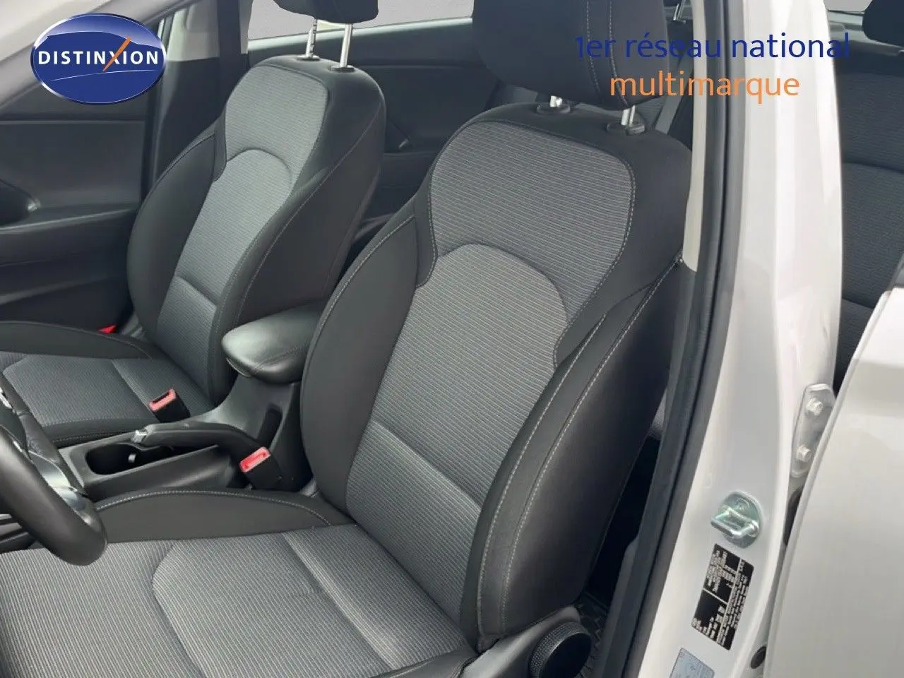 Vue intérieure côté conducteur d'une Hyundai i30 blanc nacré 2023, sièges tissu gris et noir avec accoudoir central.