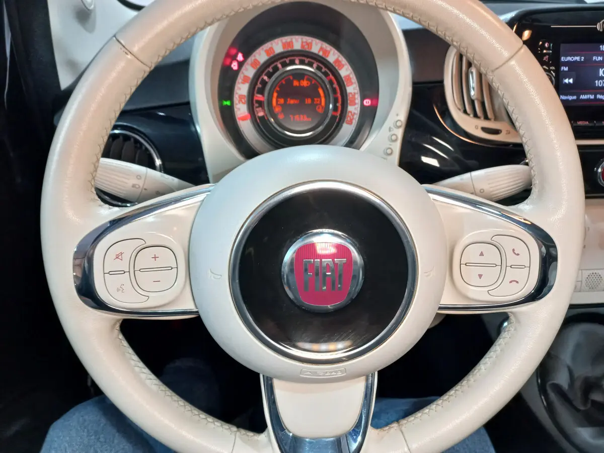 Vue rapprochée du volant beige de la Fiat 500 noire, avec commandes intégrées et tableau de bord lumineux en arrière-plan.