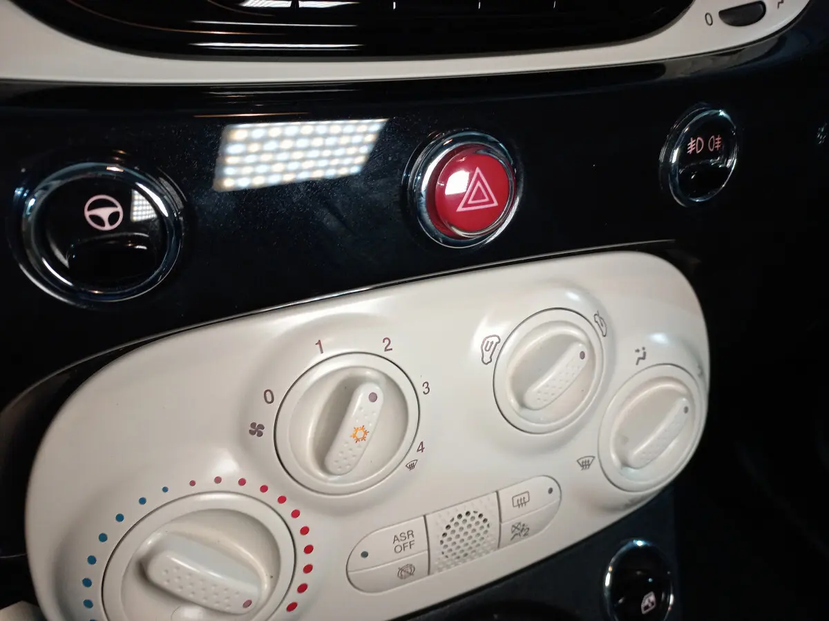 Gros plan sur les commandes de climatisation blanches et le bouton rouge de warning dans l’habitacle d’une Fiat 500 noire.