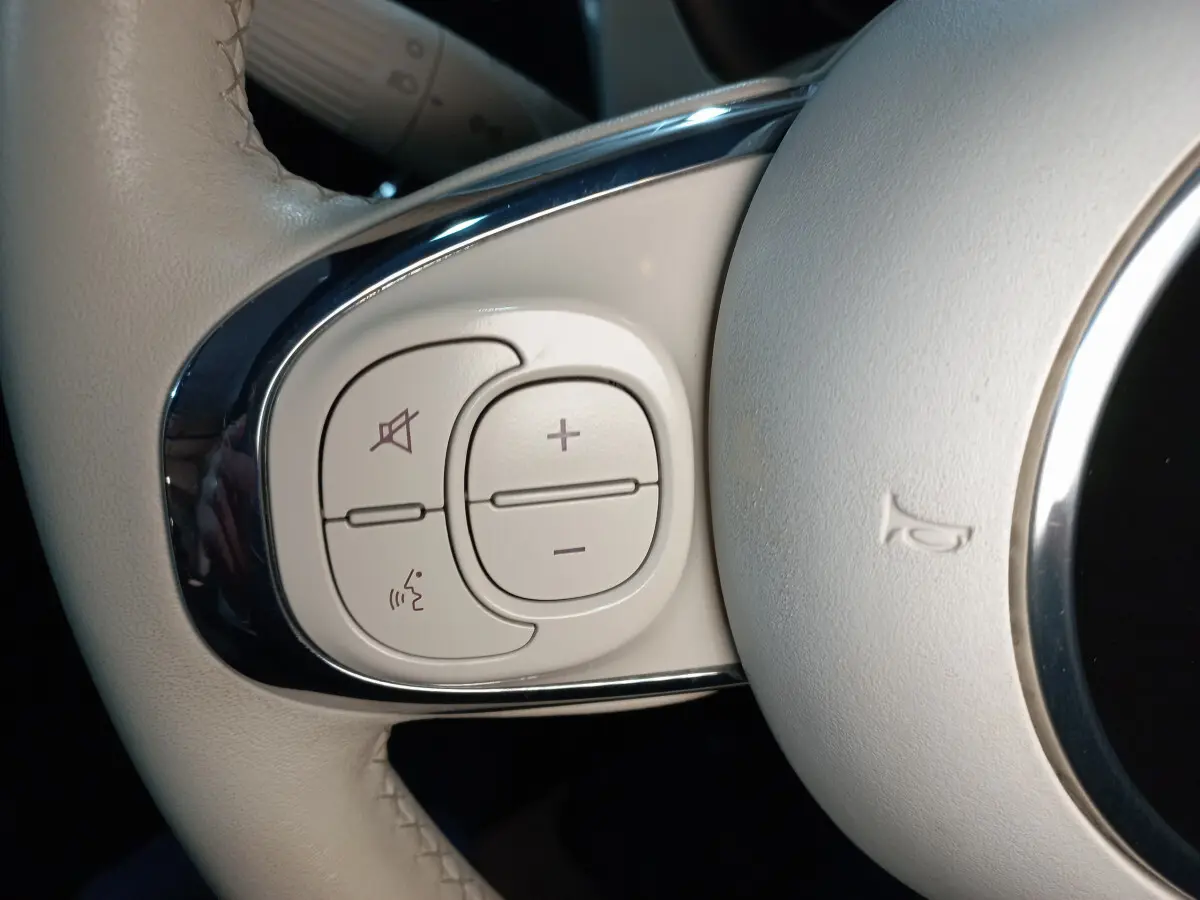 Gros plan sur les commandes de volume et de reconnaissance vocale sur le volant beige d'une Fiat 500 Lounge noire.
