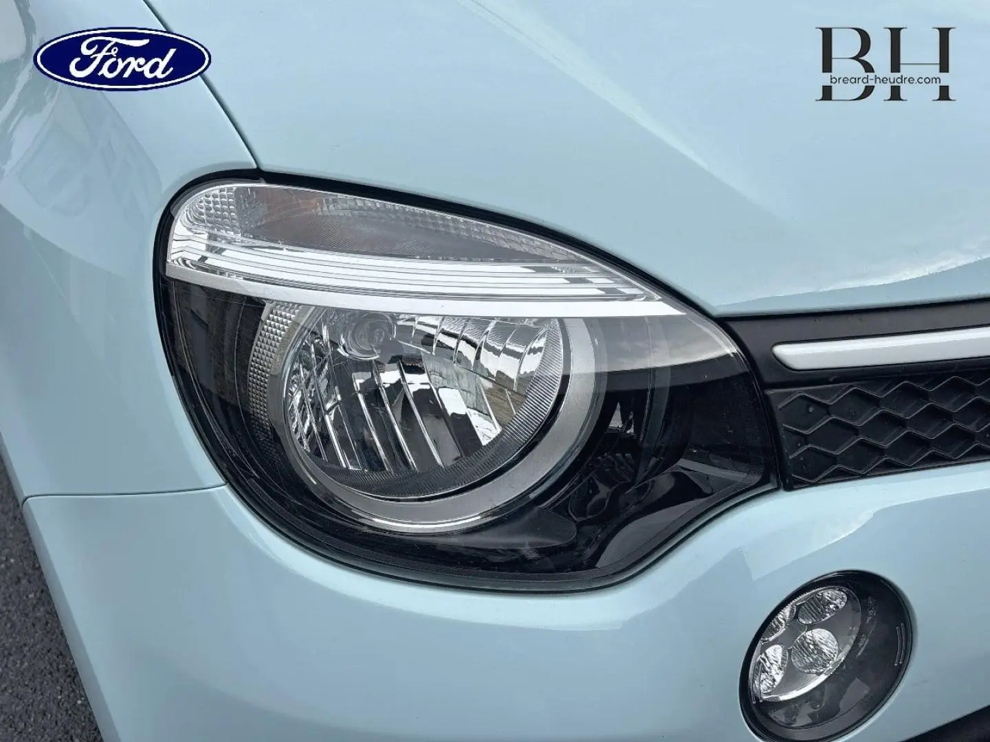 Gros plan sur le phare avant gauche d’une Renault Twingo bleu dragée avec calandre noire et logo Ford visible en reflet.