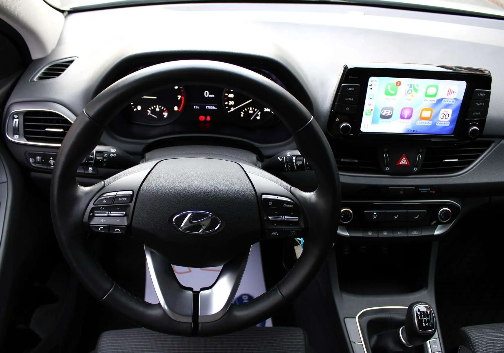 Vue intérieure centrée sur le volant et le tableau de bord d'une Hyundai i30 SW 2024 avec écran tactile et levier de vitesse manuel.