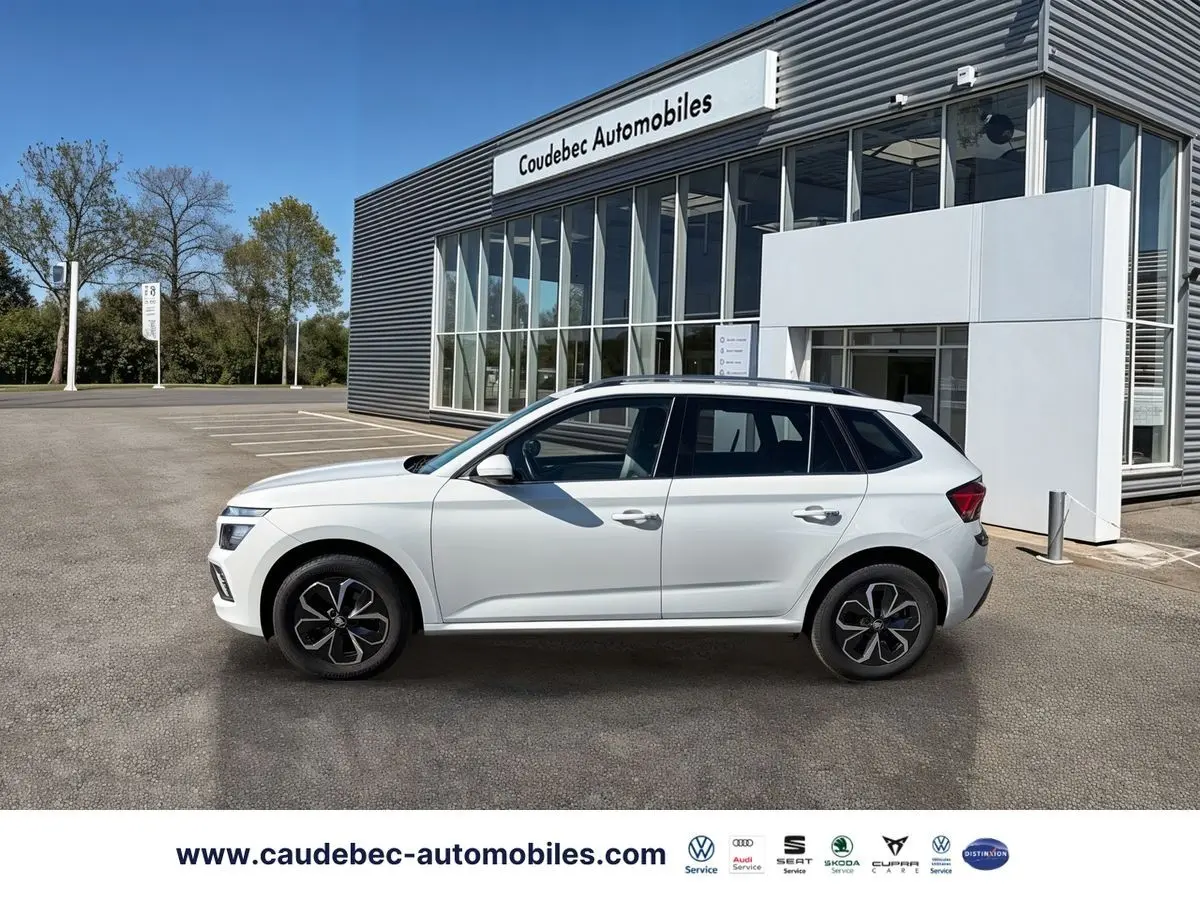 Profil côté gauche d'un Skoda Kamiq blanc 2025 garé devant un bâtiment moderne de concession automobile.