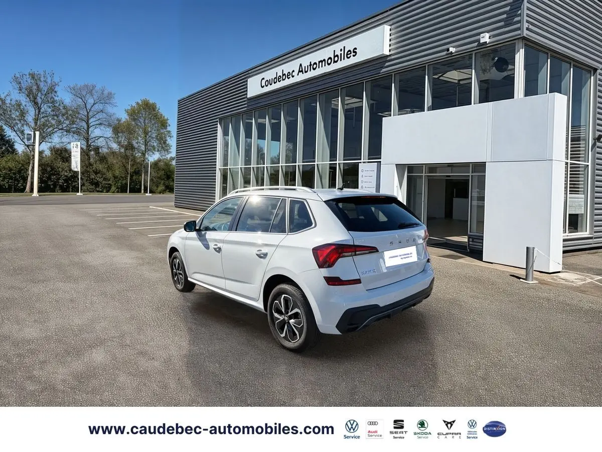 Vue 3/4 arrière droite d’un Skoda Kamiq blanc 2025 avec jantes alliage, stationné devant un bâtiment moderne.
