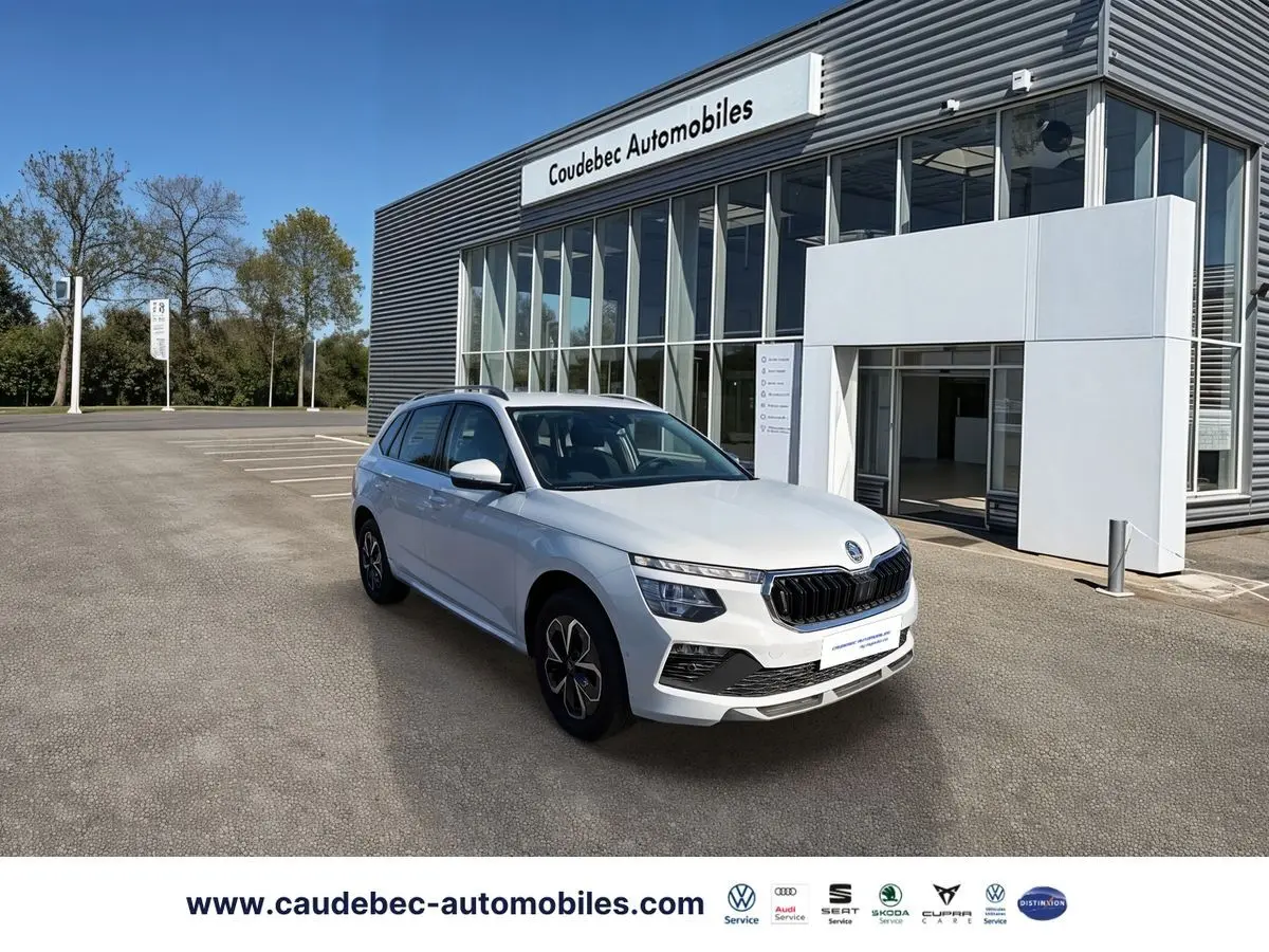 Vue 3/4 avant d'un Skoda Kamiq blanc stationné devant un bâtiment moderne de concession automobile.