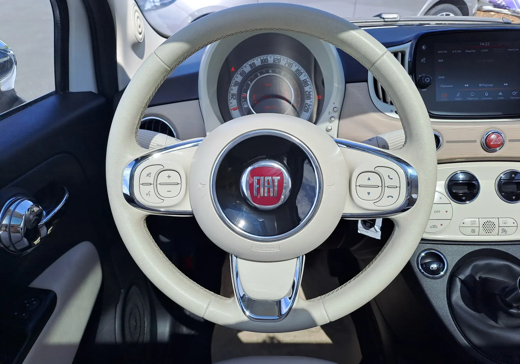 Vue frontale du volant beige clair de la Fiat 500 1.0 Hybrid 70CH DolceVita avec tableau de bord et console centrale bois clair.