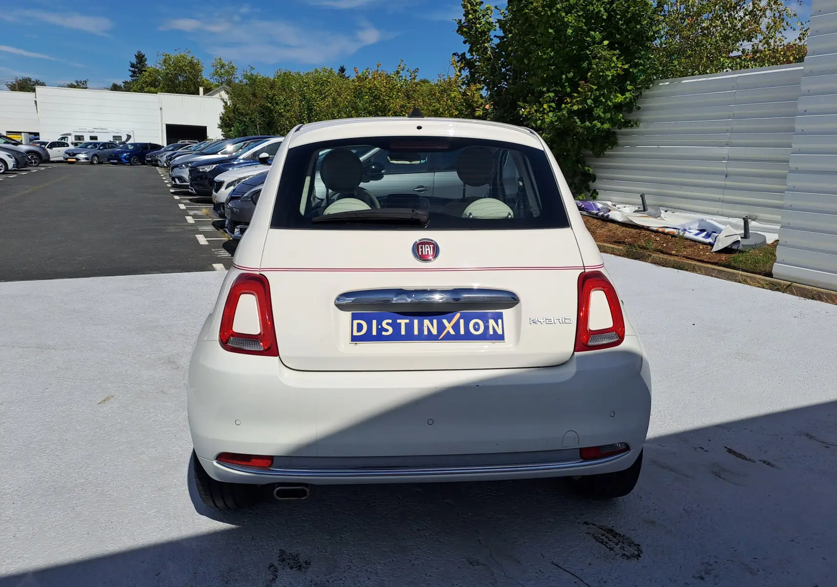 Vue arrière d'une FIAT 500 blanche hybride avec logo et plaque Distinxion, stationnée en extérieur au soleil.