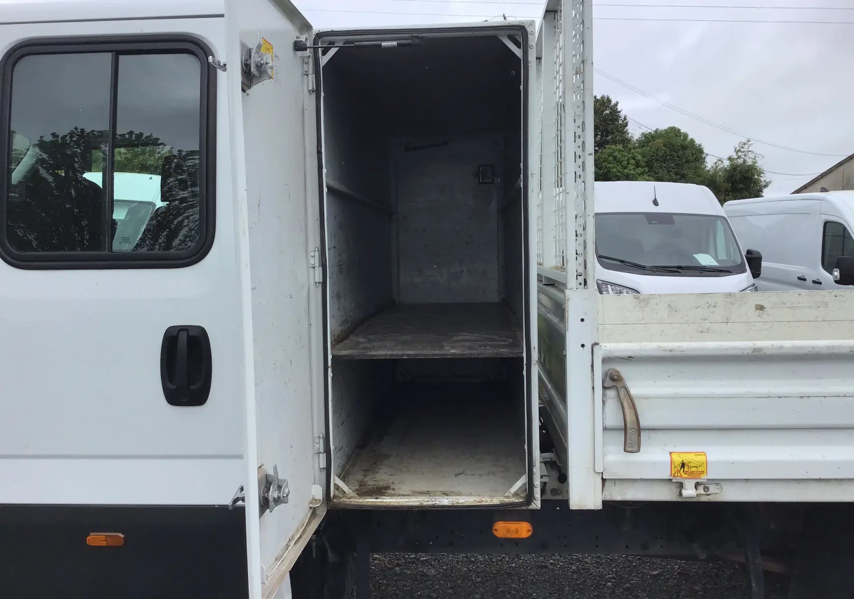 Vue latérale gauche d'un Iveco Daily blanc avec benne et coffre ouvert montrant l'intérieur vide du rangement.
