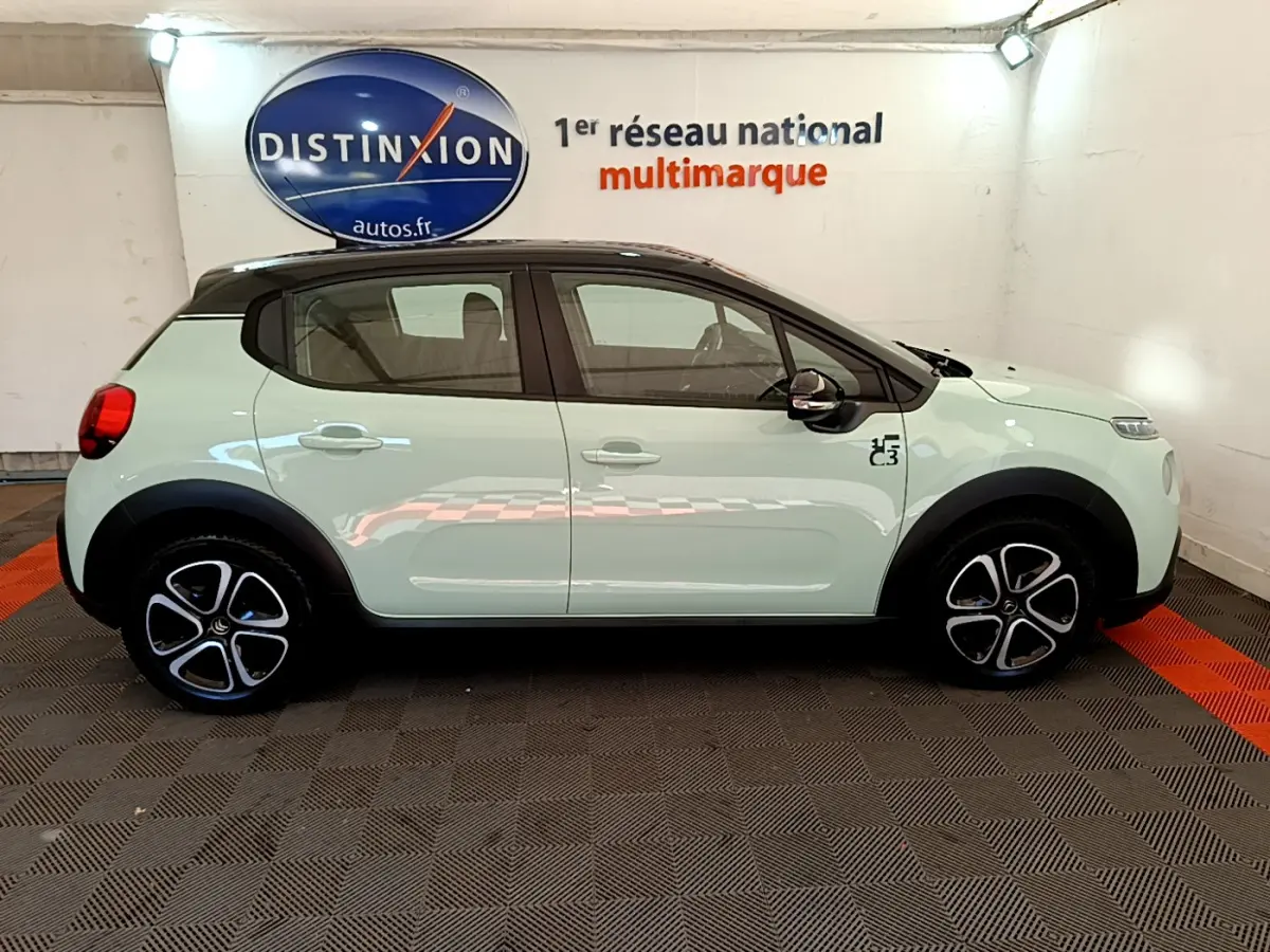 Profil côté gauche d'une Citroën C3 PureTech 82 vert clair avec toit noir et jantes stylisées en intérieur showroom.