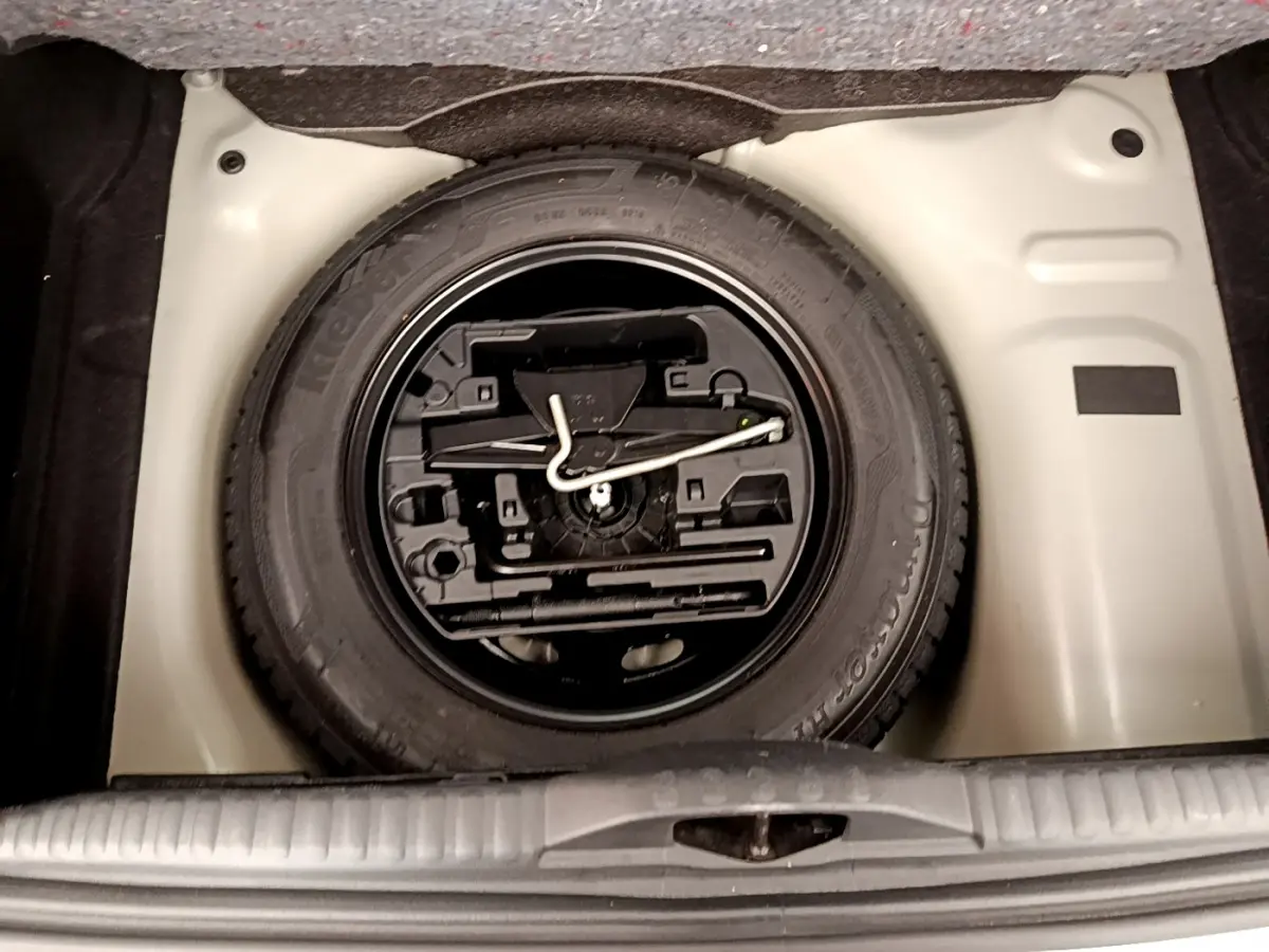 Vue plongeante sur la roue de secours noire et son kit d'outils dans le coffre beige d'une Citroën C3 verte.