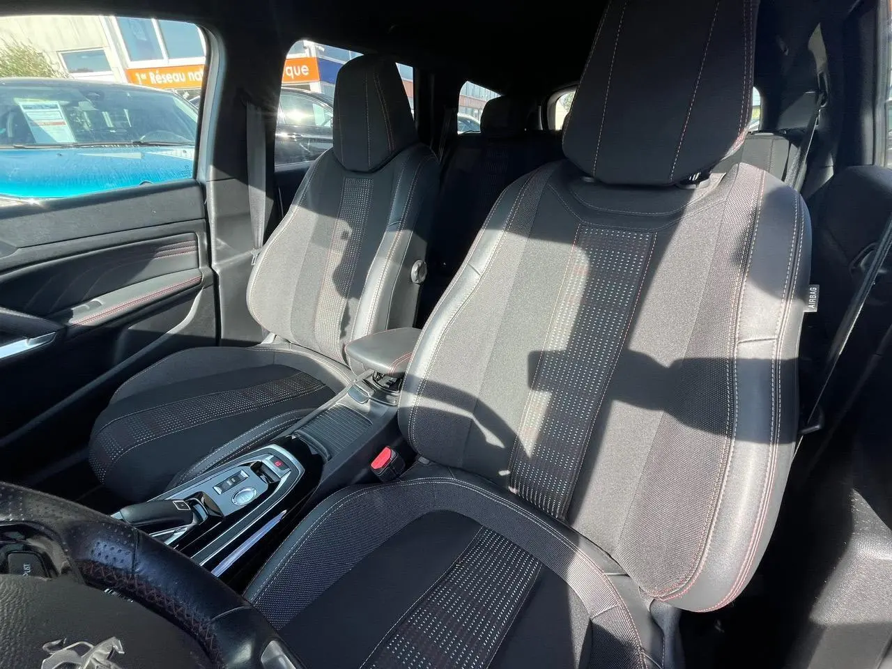 Vue intérieure côté avant droit des sièges en tissu noir avec surpiqûres rouges et console centrale d'une Peugeot 308 SW gris clair.