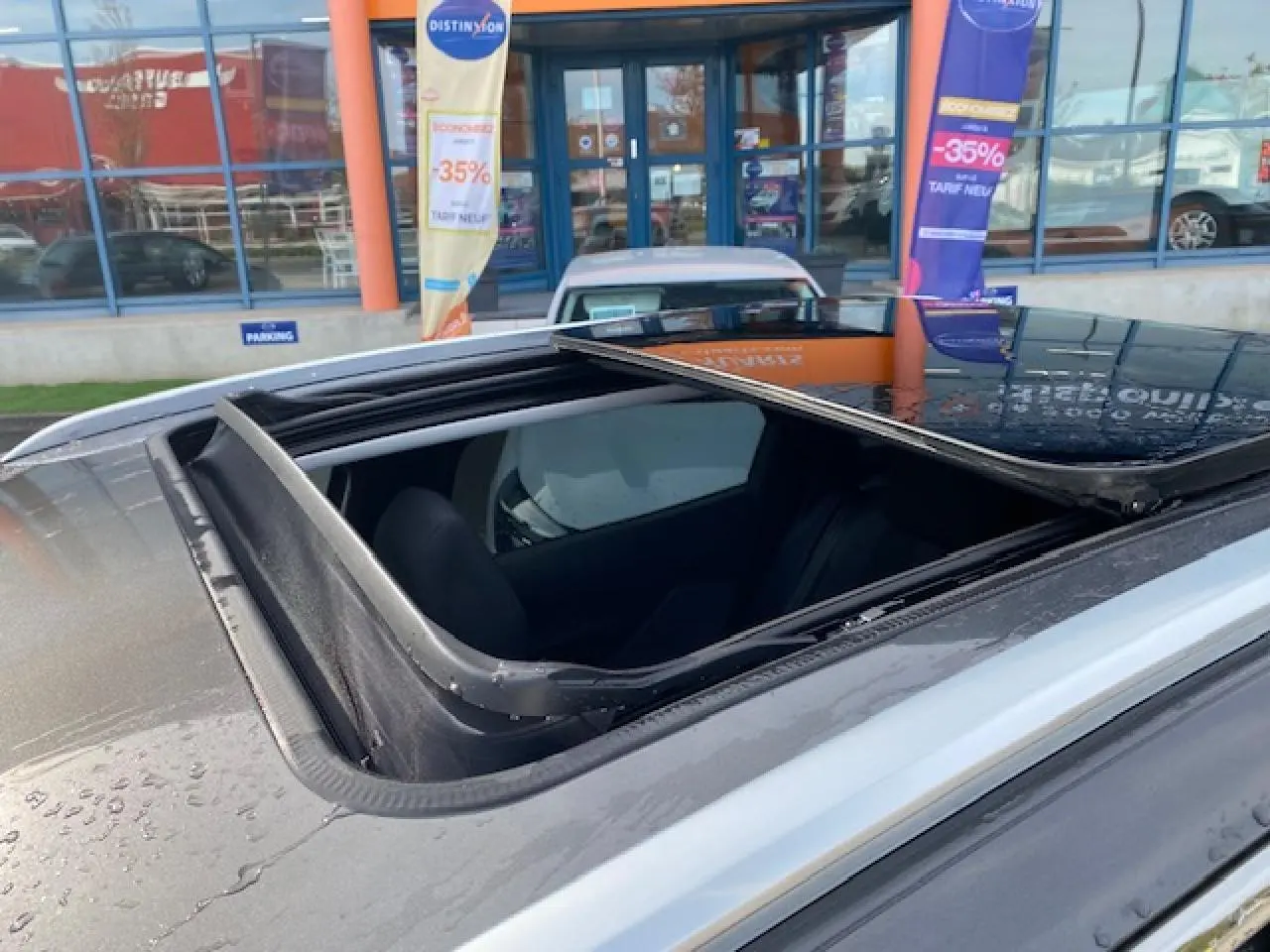 Toit ouvrant panoramique ouvert vu en gros plan sur un Peugeot 3008 gris platinium stationné devant une vitrine.