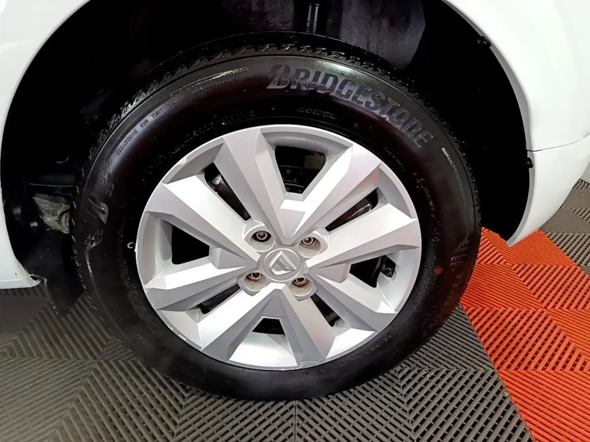 Gros plan sur la roue avant droite d'une Dacia Sandero blanche, pneu Bridgestone visible, sur sol à damiers gris et orange.