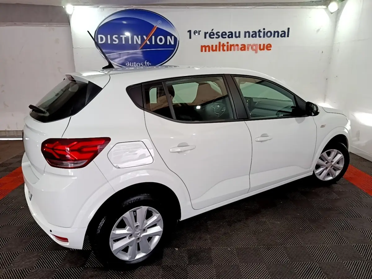 Vue 3/4 arrière droite d'une Dacia Sandero blanche 2022 dans un showroom avec logo Distinxion en arrière-plan.