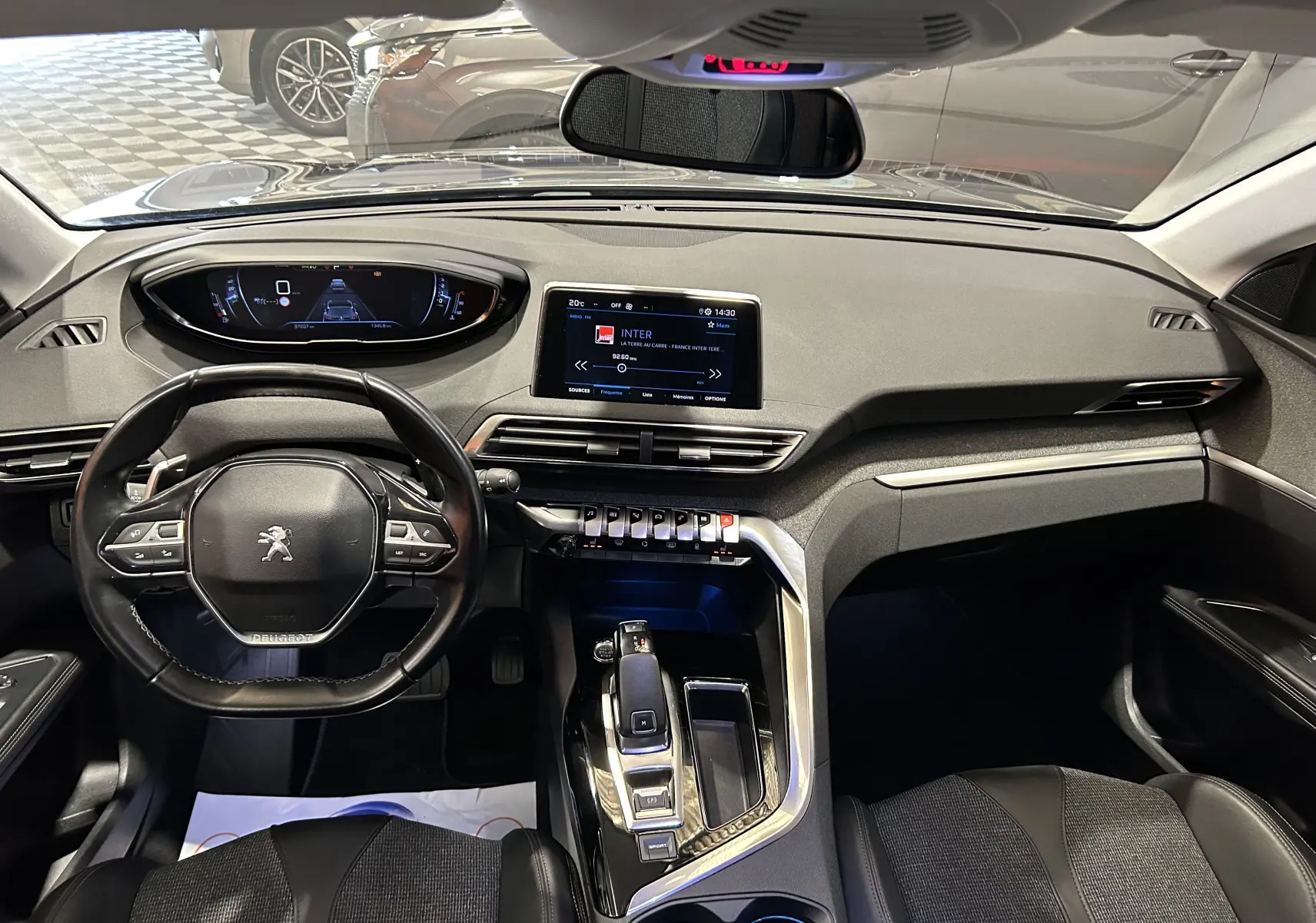 Vue intérieure frontale du tableau de bord et volant noir du Peugeot 5008 gris, avec écran tactile central et levier de boîte automatique.