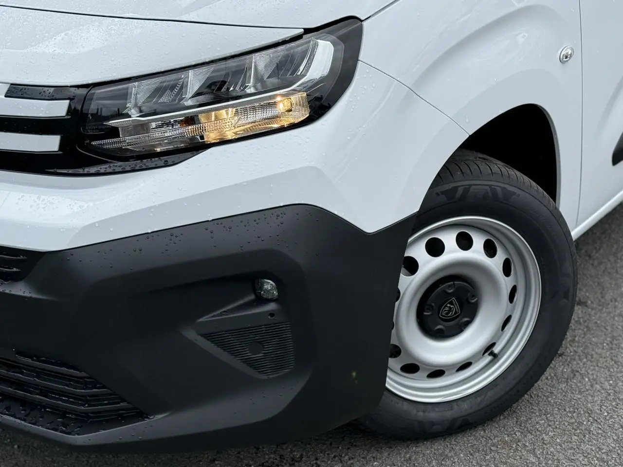 Gros plan sur l'avant droit blanc du Peugeot Partner XL 2025, mettant en valeur le phare allumé et la roue acier.