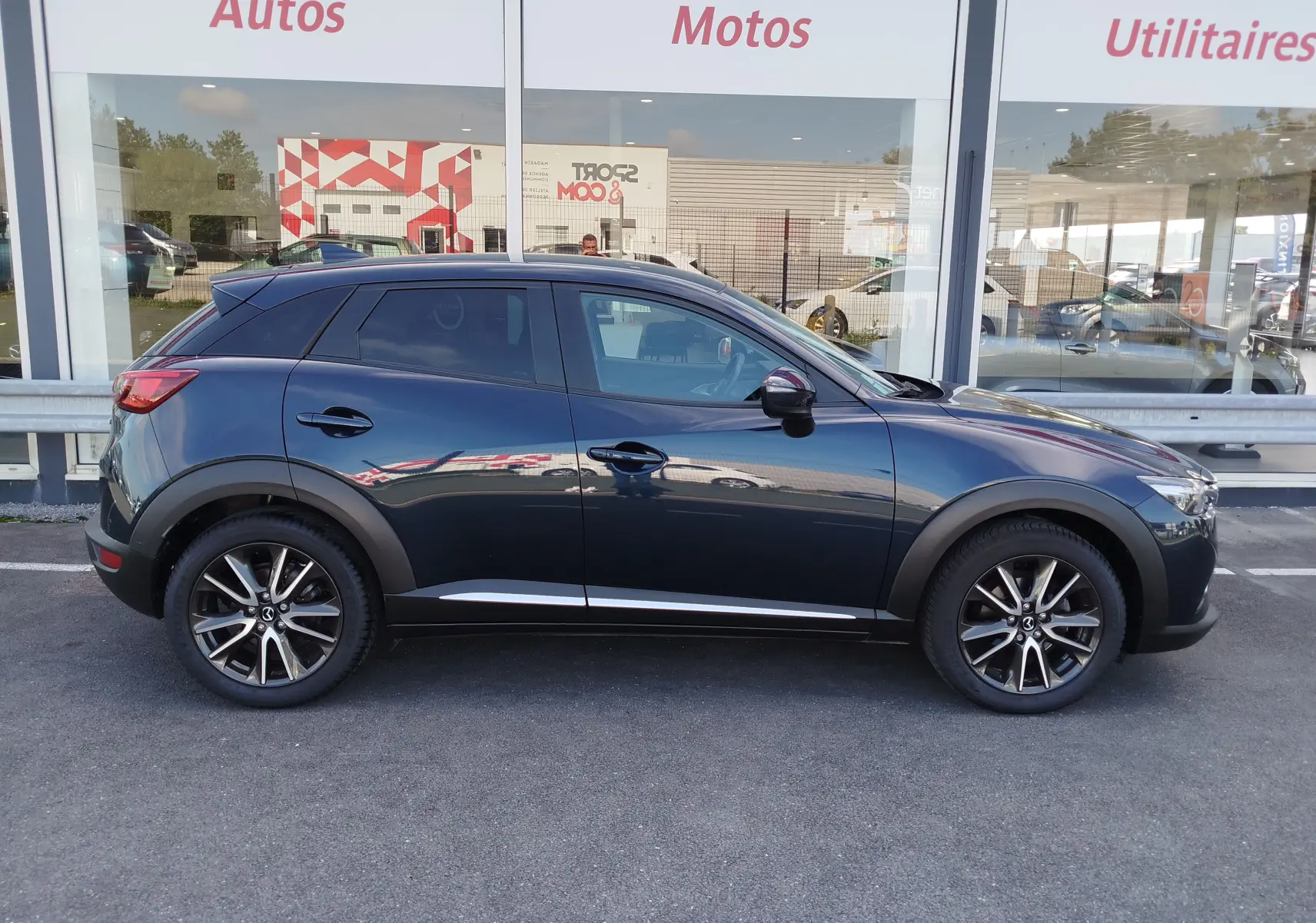 Profil côté gauche d'un Mazda CX-3 2016 bleu foncé avec jantes alliage et vitres teintées, stationné devant une vitrine.