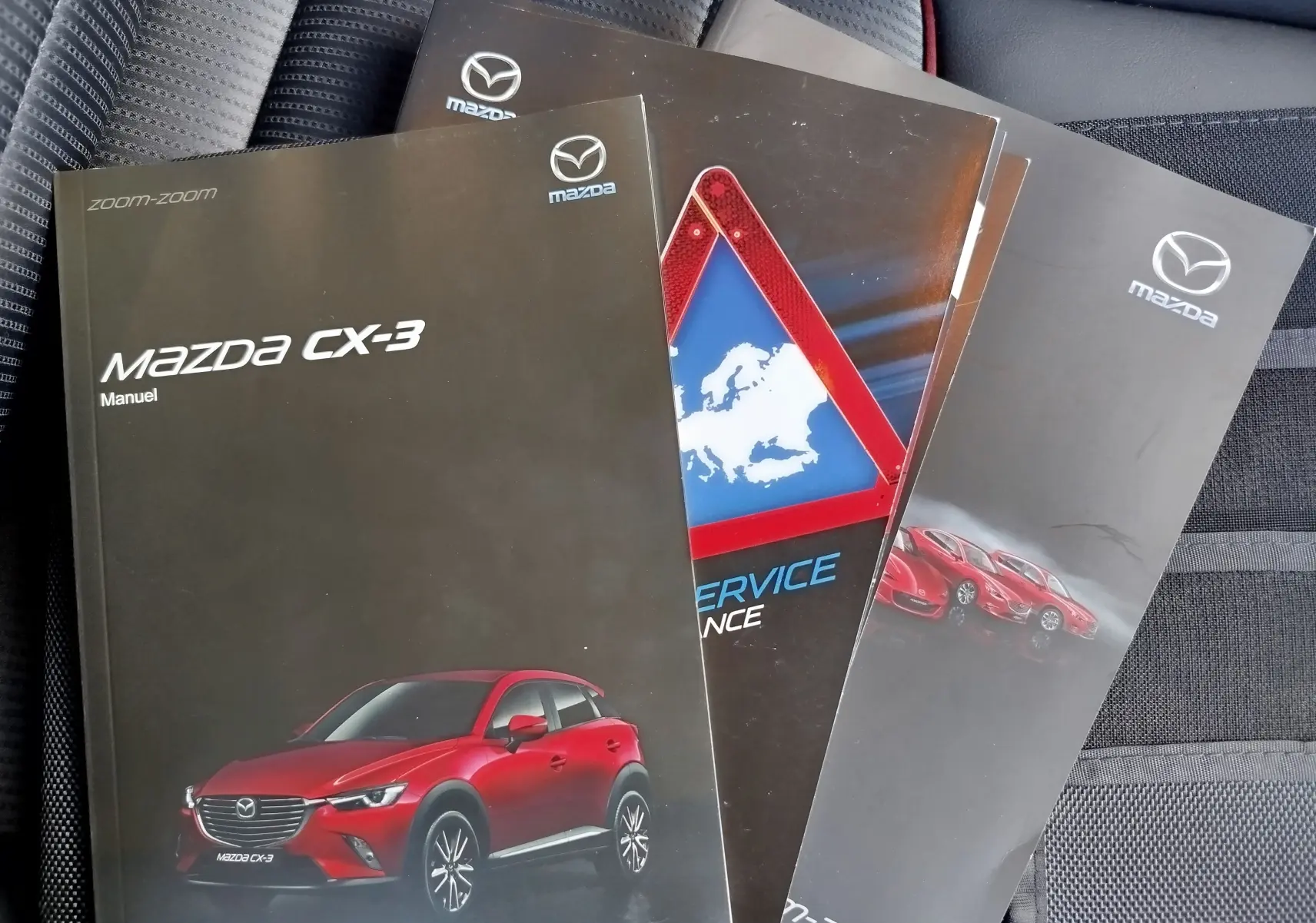 Manuel Mazda CX-3 rouge en vue de profil posé sur un siège auto gris texturé, avec autres brochures Mazda visibles.
