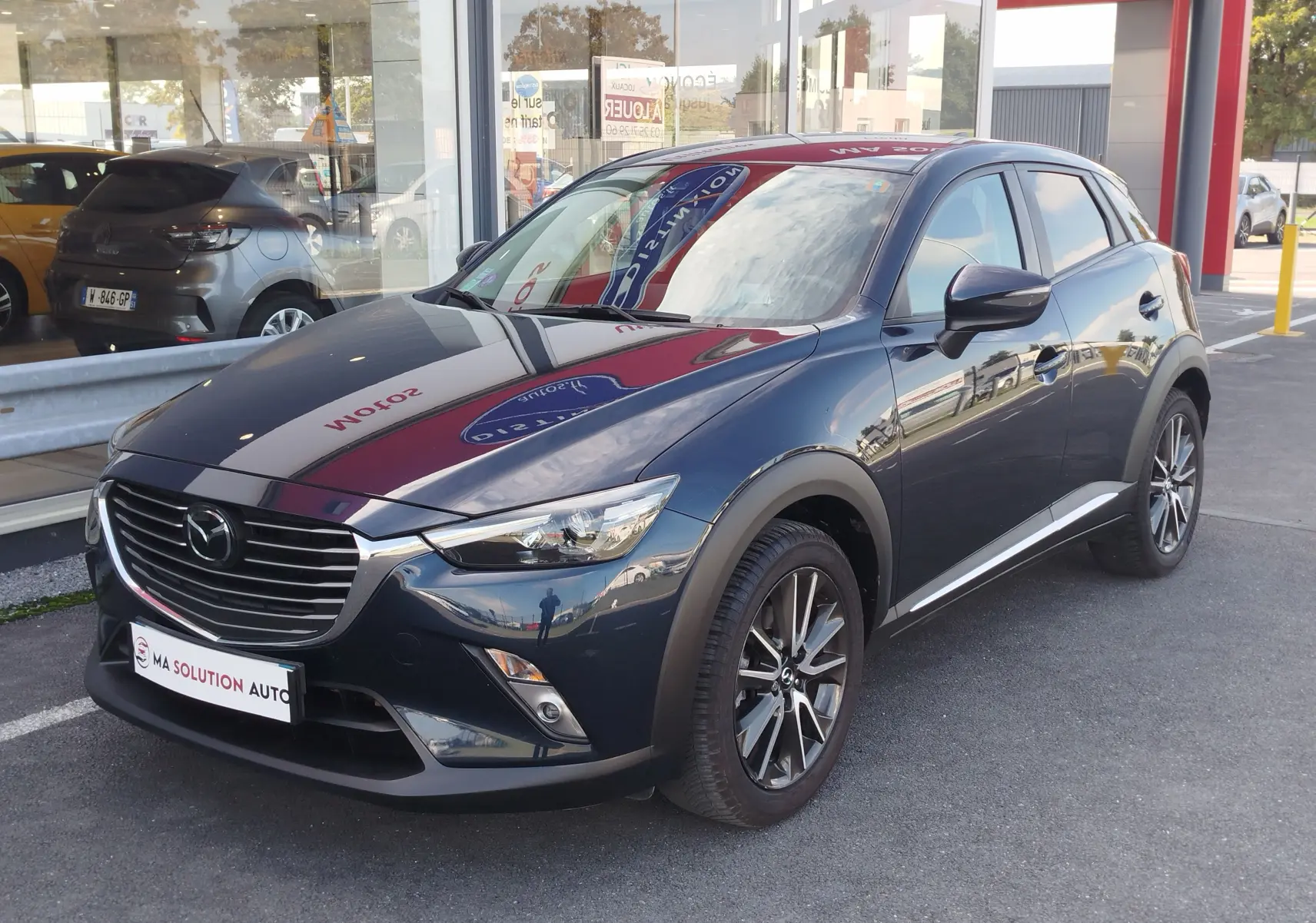 Vue 3/4 avant d'un Mazda CX-3 bleu foncé avec jantes alliage et calandre chromée, stationné devant un showroom.