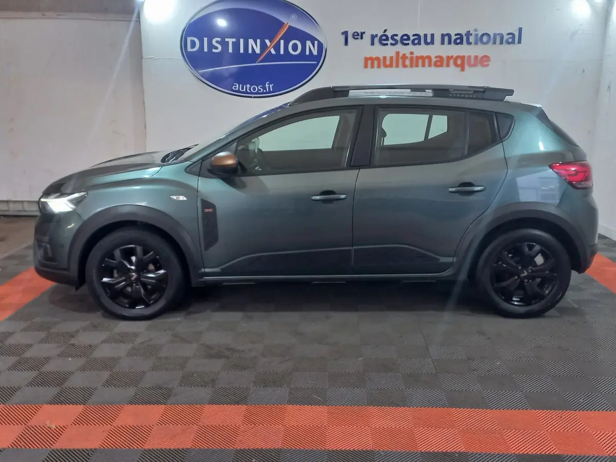 Vue de profil côté gauche d'une Dacia Sandero Extreme+ TCe 110 2024 vert foncé avec jantes noires et barres de toit.