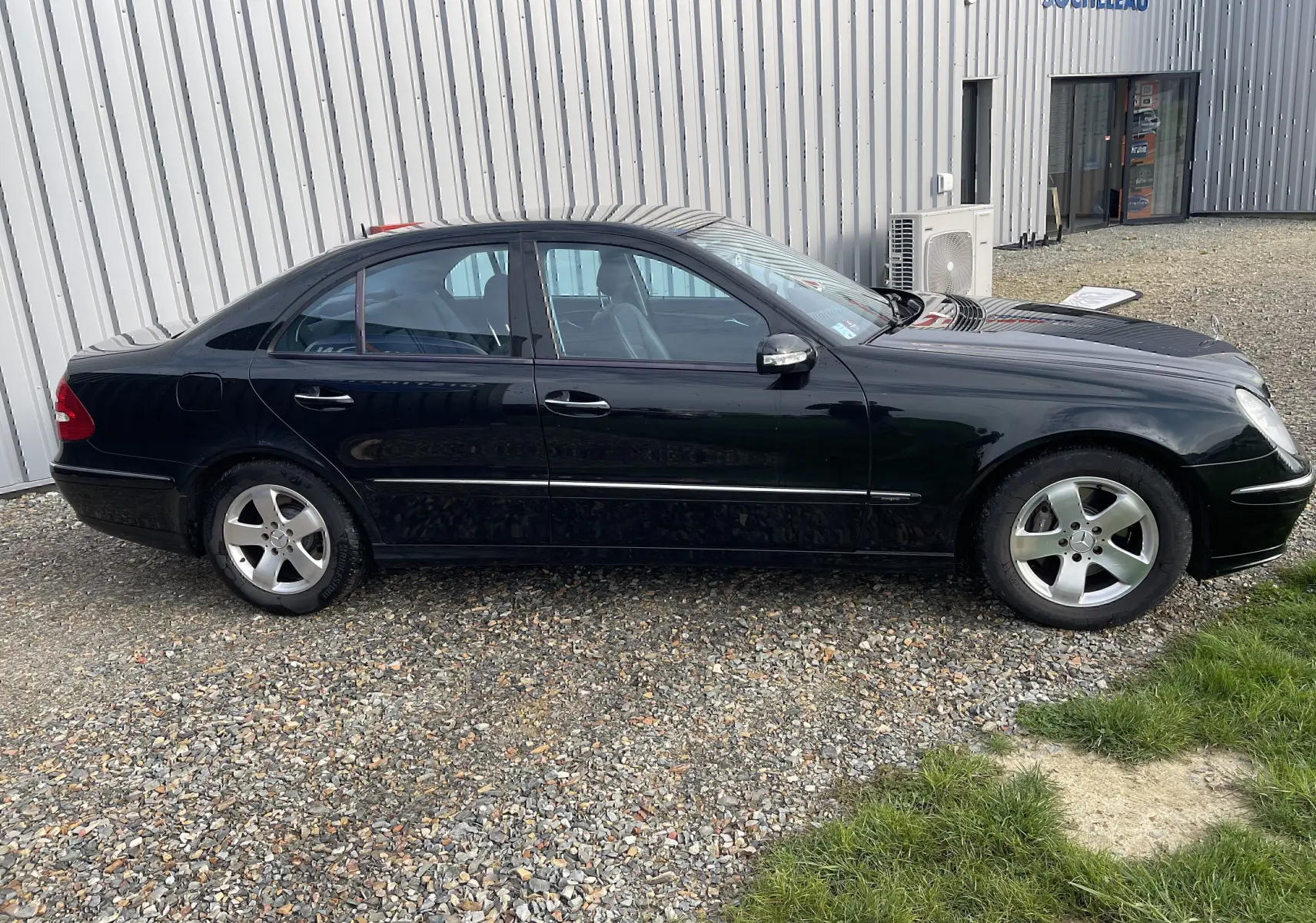 Vue de profil côté gauche d'une Mercedes Classe E 320 CDI Elegance noire de 2003, garée sur un sol gravillonné.