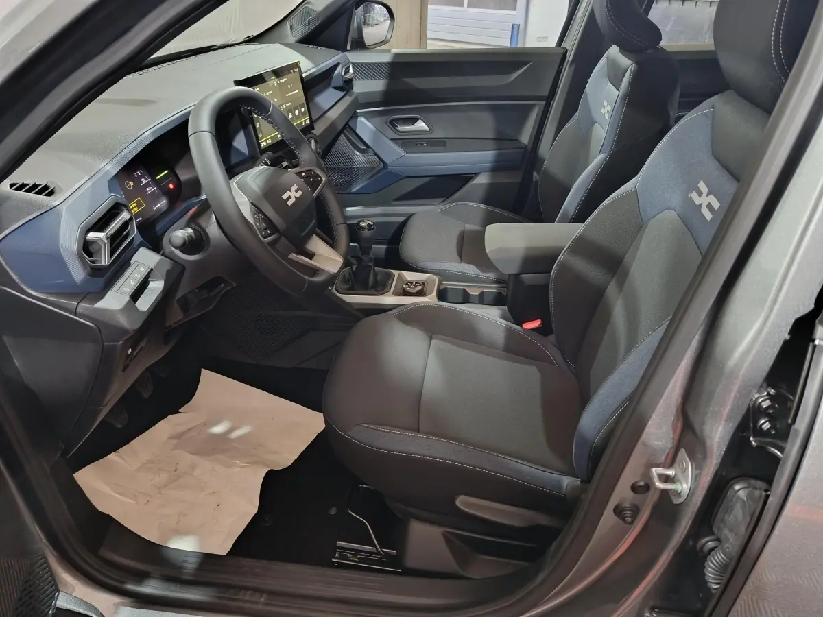 Vue intérieure côté conducteur du Dacia Bigster gris foncé, montrant le tableau de bord moderne et les sièges tissu avec logo.
