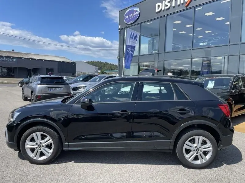 Audi Q2 TFSI 150CV noir métal vu de profil côté gauche, garé devant un showroom avec plusieurs autres véhicules.