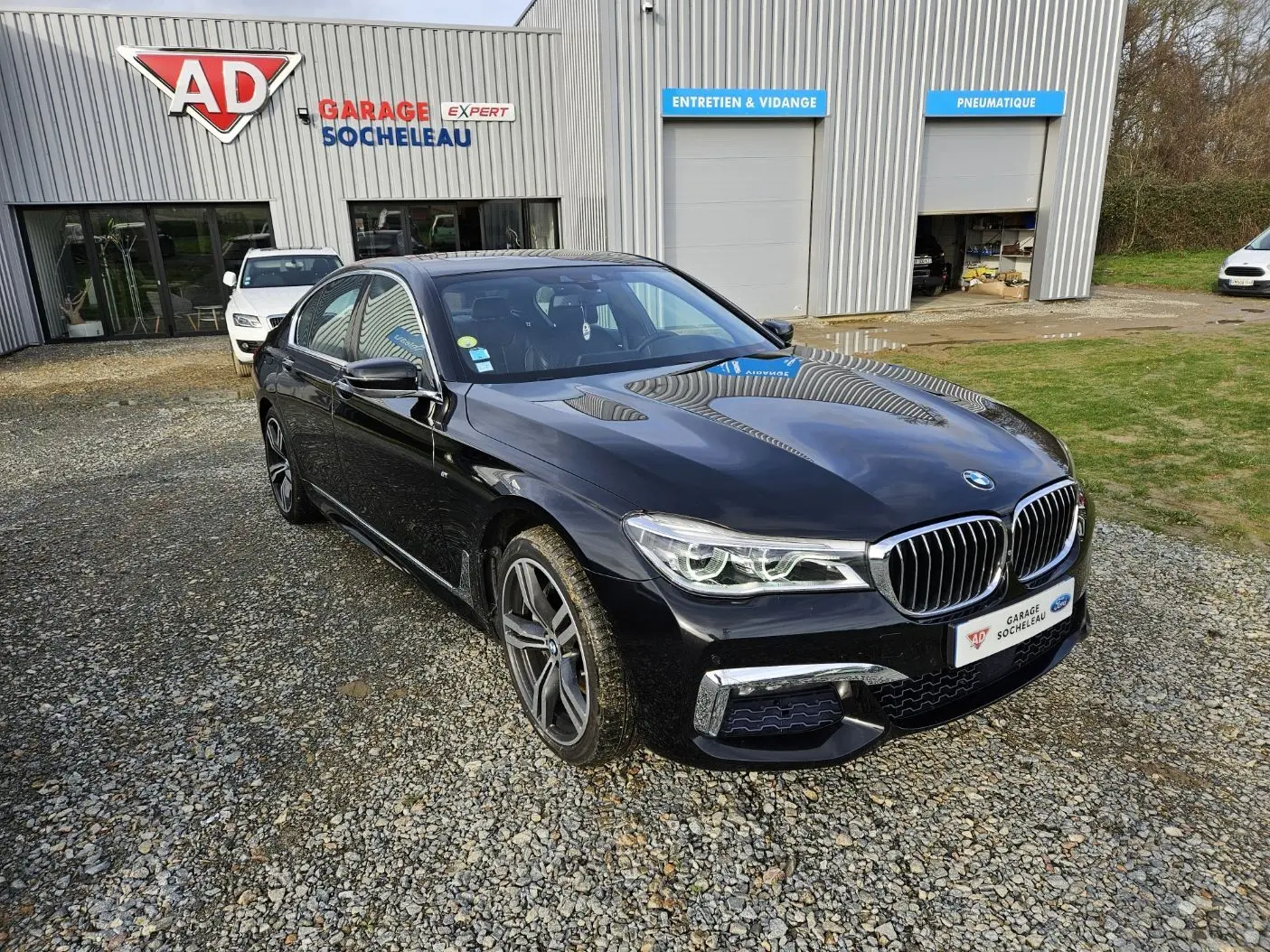 BMW Série 7 G11 noire vue en 3/4 avant droit, avec calandre chromée et jantes sport devant un garage AD Socheleau.