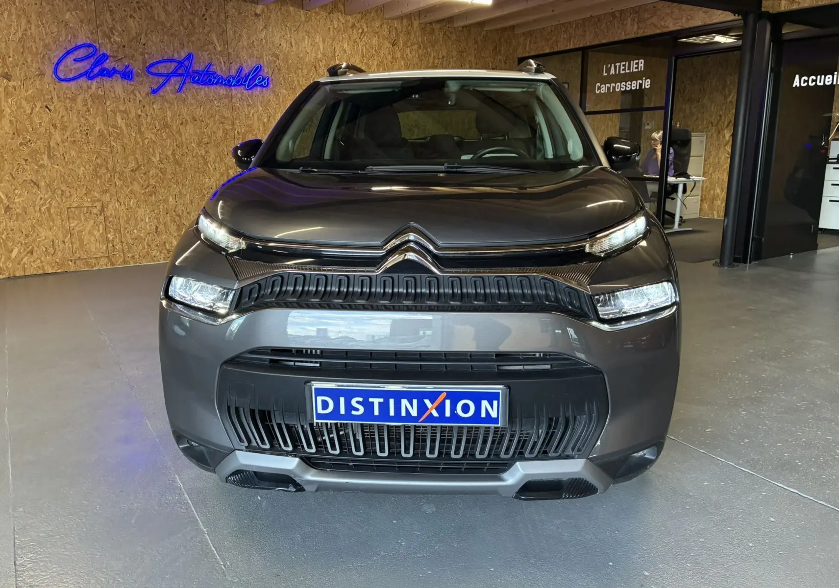 Vue frontale d'un Citroën C3 Aircross gris platinium métal avec calandre noire et plaques Distinxion en intérieur showroom.