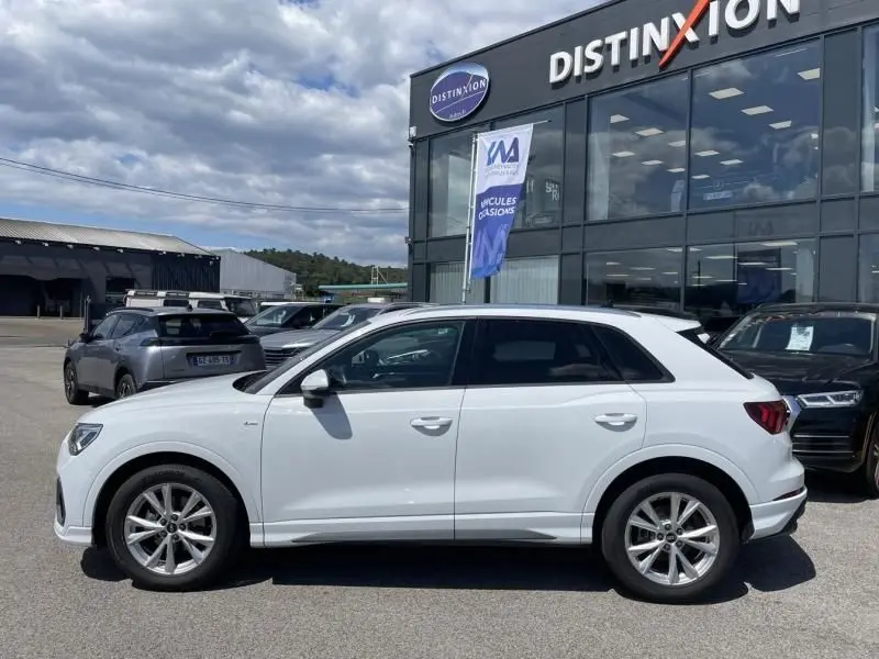 Audi Q3 blanc vue de profil côté gauche, finition S-Line avec jantes alliage, stationné devant un concessionnaire Distinxion.
