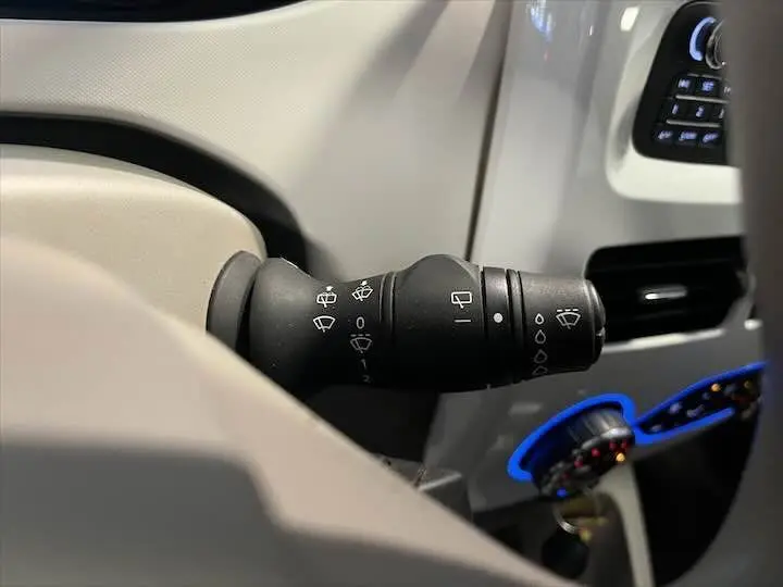 Gros plan sur la manette d'essuie-glace noire dans l'habitacle clair d'une Renault ZOE blanc glacier, avec commandes lumineuses bleues.