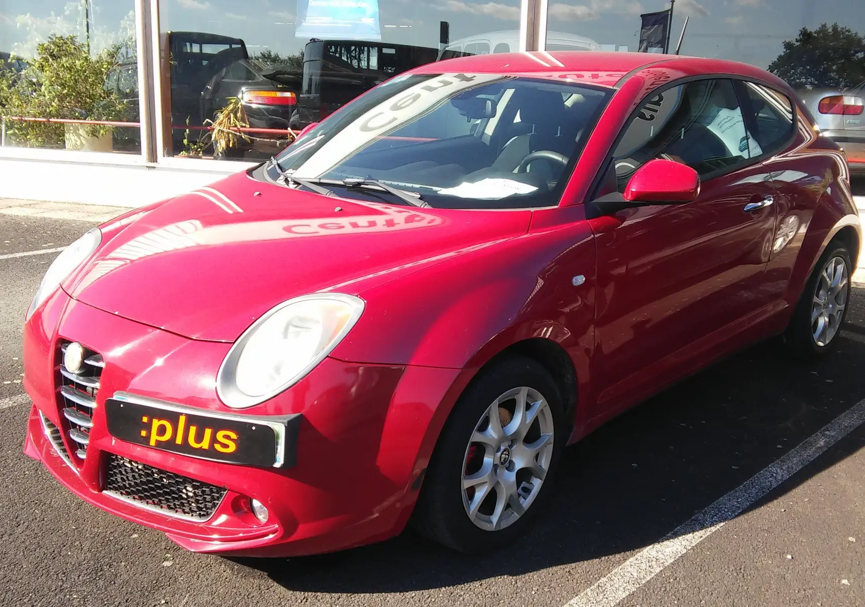 Alfa Romeo MiTo rouge vue 3/4 avant droit, avec jantes alliage et calandre distinctive sur parking.