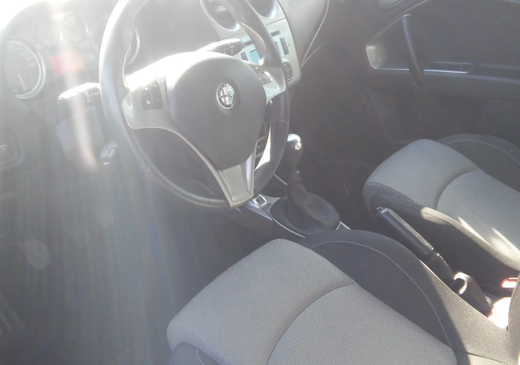 Intérieur avant droit de l'Alfa Romeo MiTo 2010, volant cuir avec logo, sièges tissu gris et levier de vitesse manuel.
