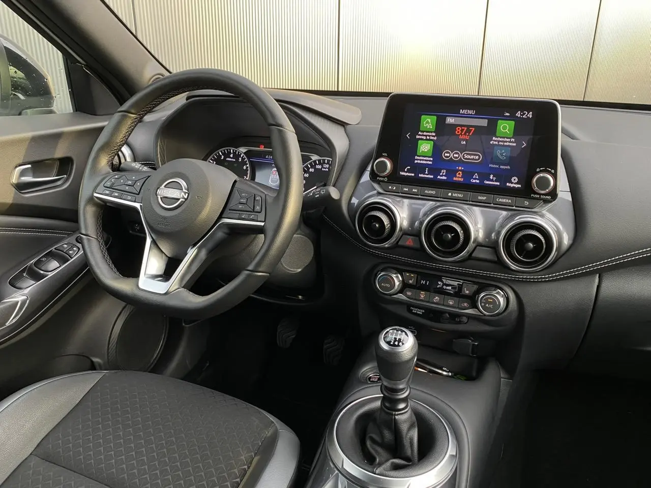Vue intérieure côté conducteur du Nissan Juke blanc perle 2024 avec volant cuir, écran tactile et boîte manuelle.